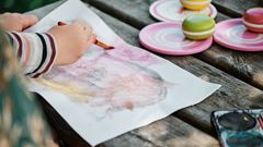 Ett barn sitter vid ett bord utomhus och målar med vattenfärg.