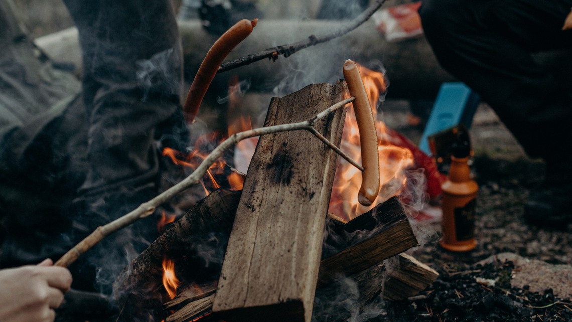 Några personer sitter runt en eld och grillar korv i skogen.